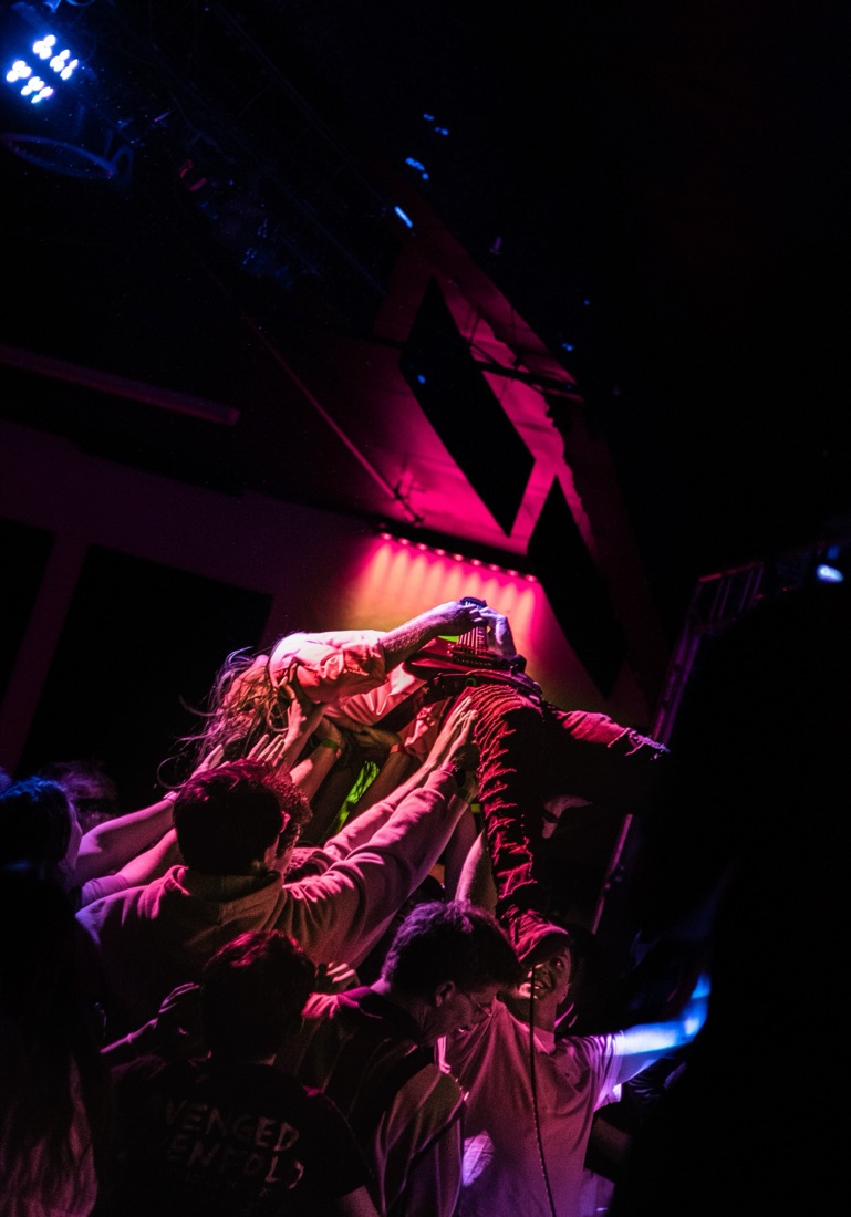 Guitarist crowd surfing while playing, held up by the crowd in pink light