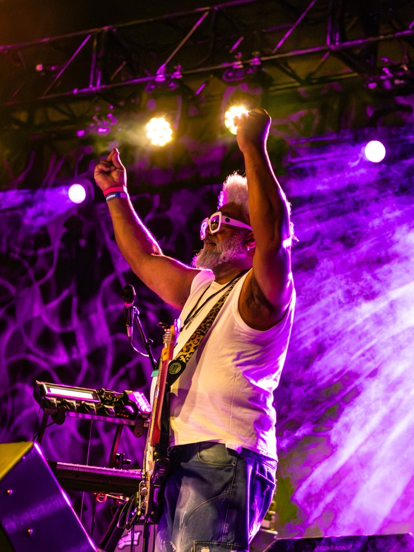 Guitarist in white glasses with both fists raised under purple lights