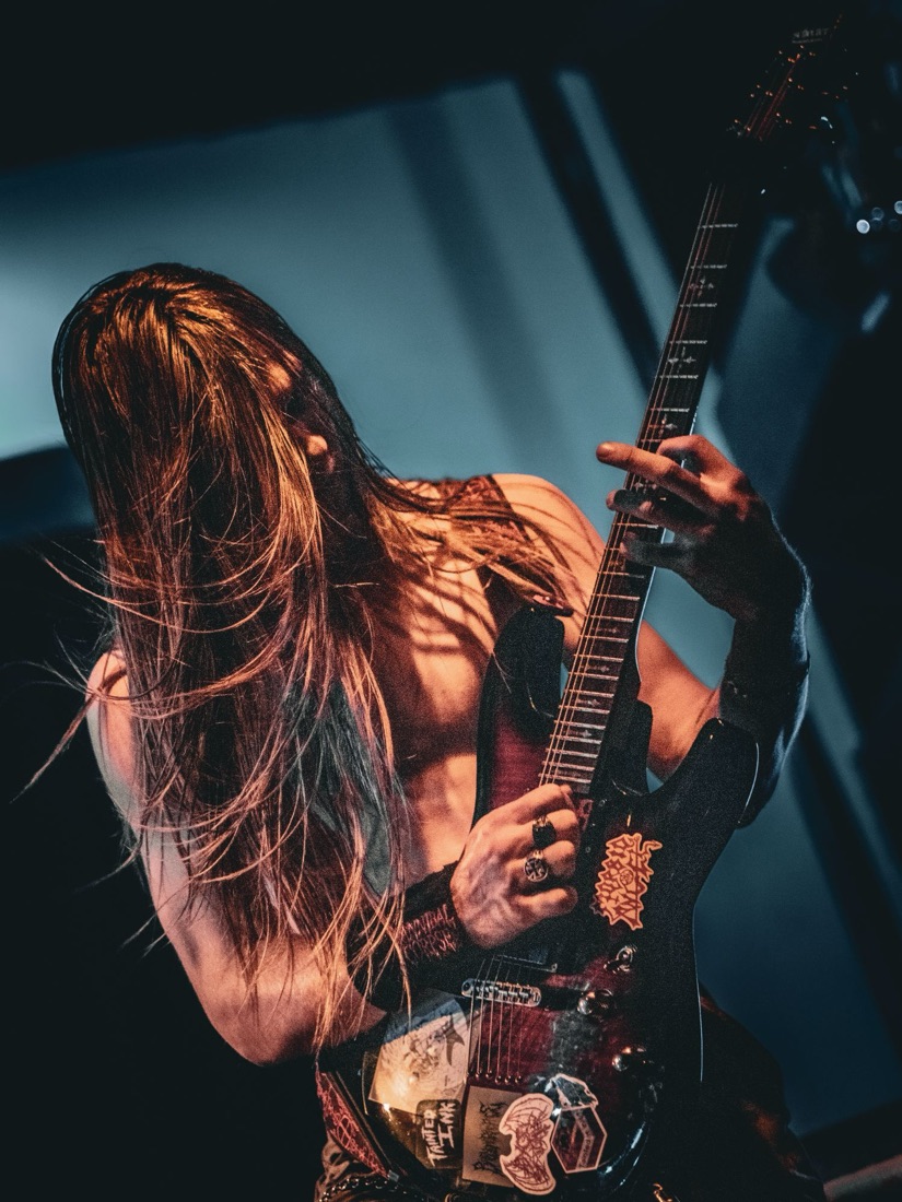 Shirtless guitarist with long hair over his face, warm light against blue