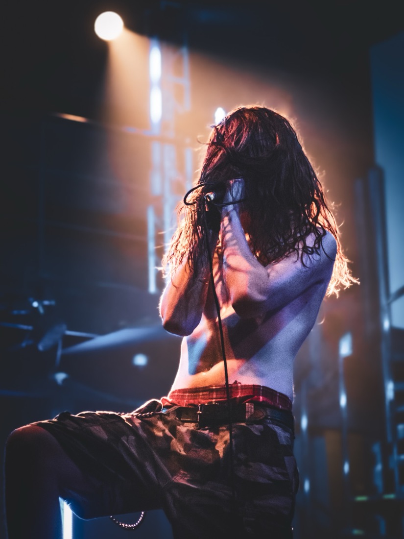 Shirtless vocalist backlit by a warm spotlight, hair covering his face