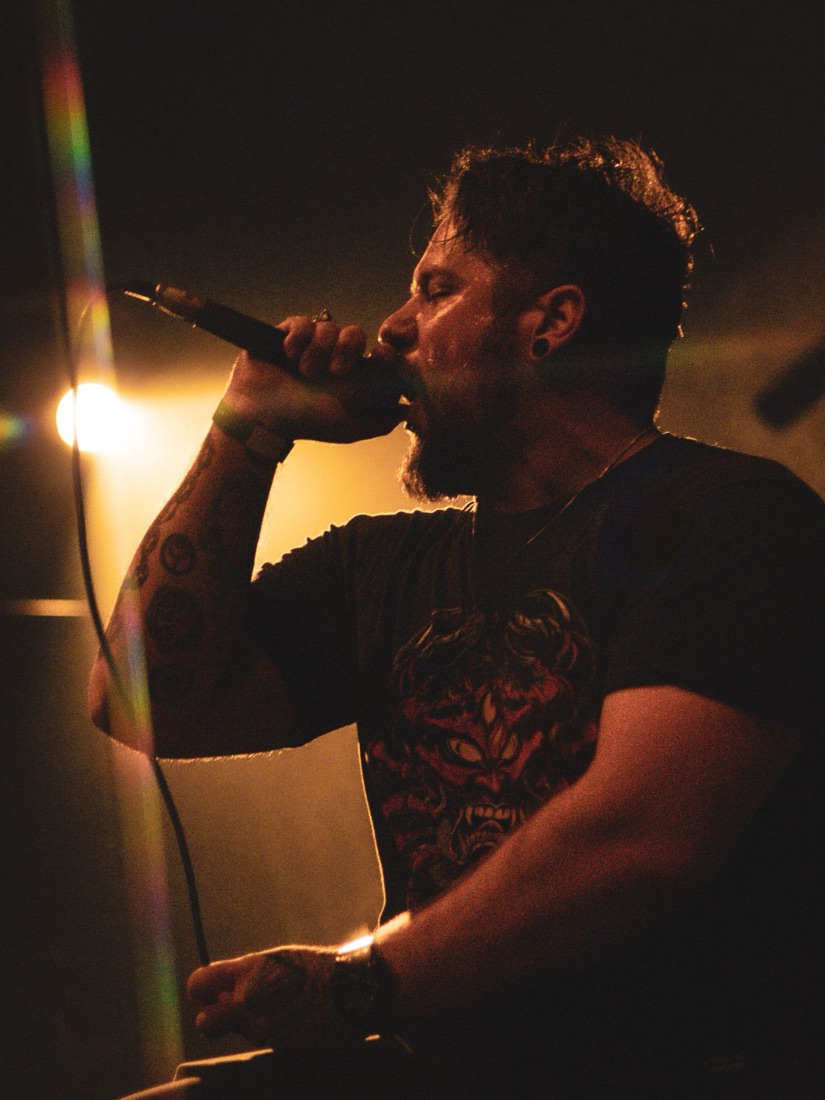 Bearded vocalist singing into a microphone, backlit in warm light